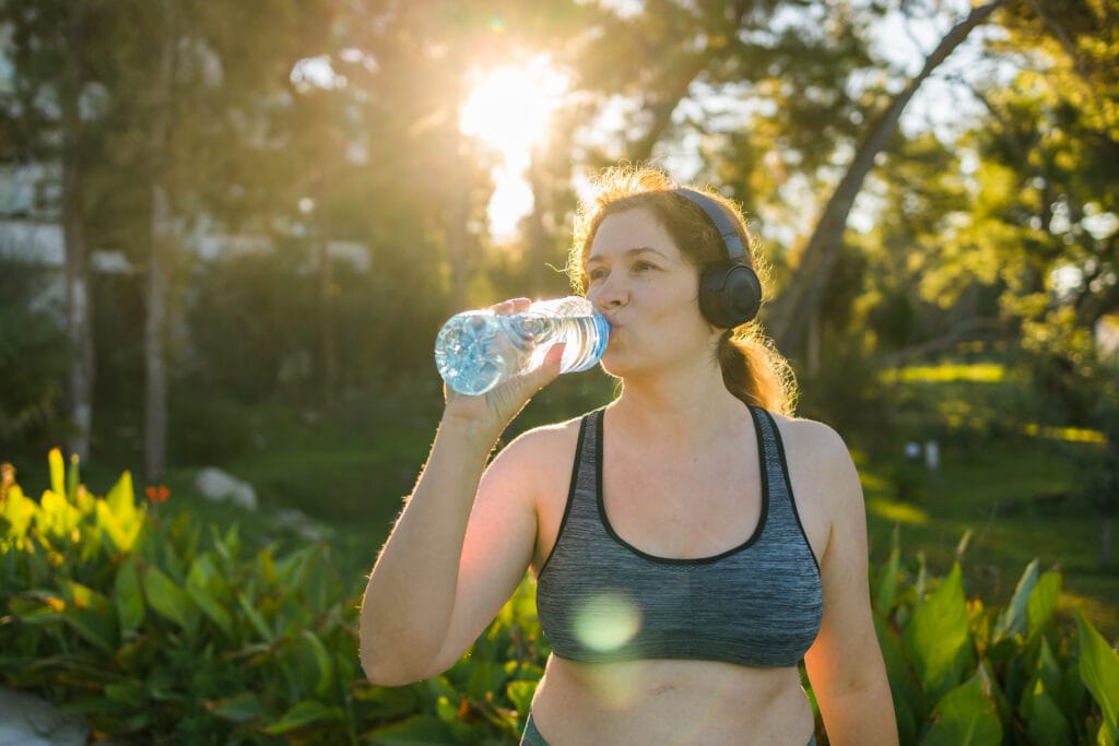 Overweight woman drinking water after jogging in the park. Portrait of young plus-size thirsty woman with a bottle of water outdoors copy space. Sports healthcare and weight loosing fitness and well-being concept