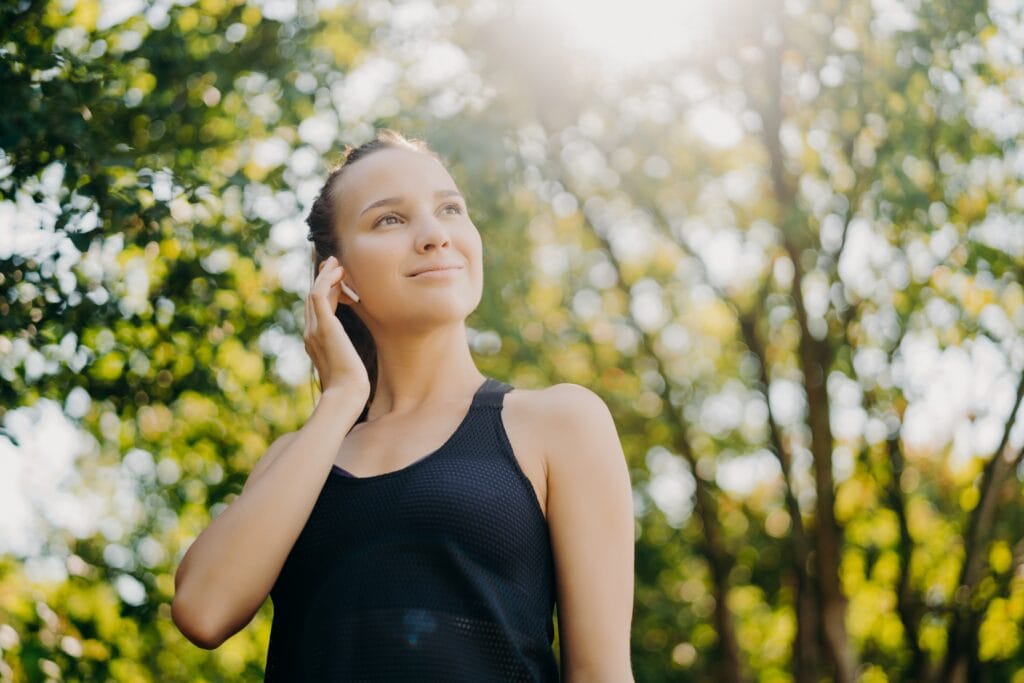 Outdoor shot of pleased athletic woman focused into distance stays active leads healthy lifestyle wears sportsclothes poses against blurred nature background. Sunny day is good for workout outside