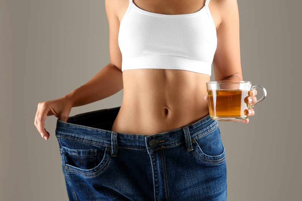 Young woman in old big jeans with cup of tea showing her diet results on beige background, closeup